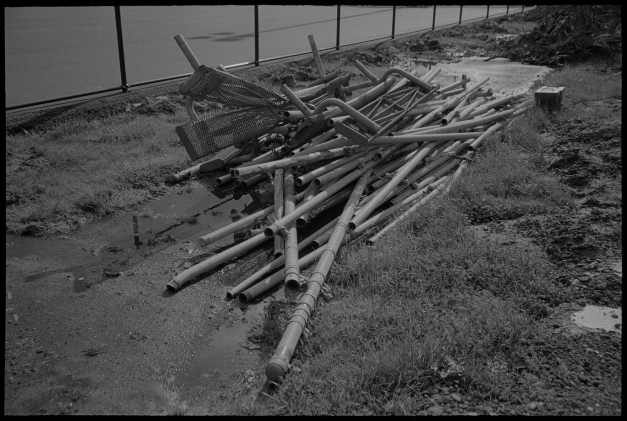 The same negative inverted into a black and white positive: a pile of grey metal tubes and a bent mesh bench dumped on muddy grass beside a railing, a road and water behind, with a date stamp in the lower right corner.